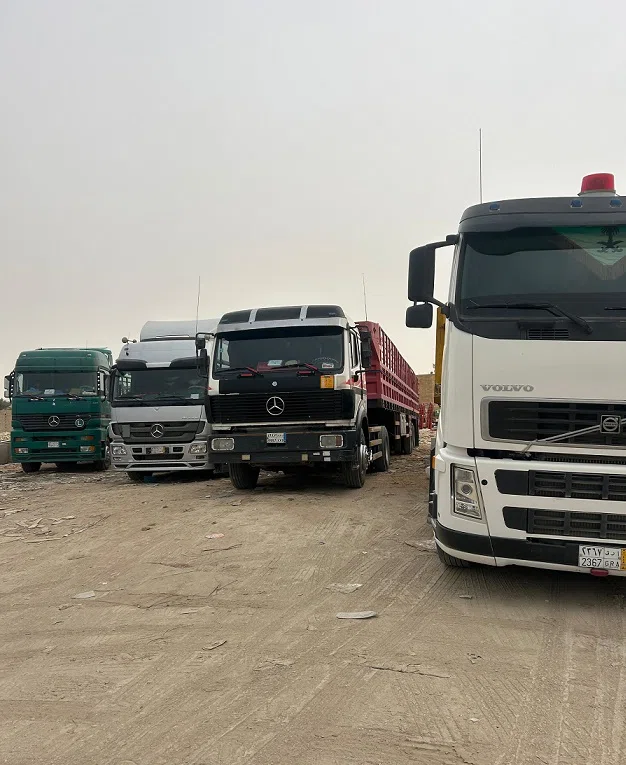 مجموعة شاحنات نقل ثقيل متوقفة في ساحة ترابية، تشمل شاحنات فولفو ومرسيدس مخصصة لنقل البضائع