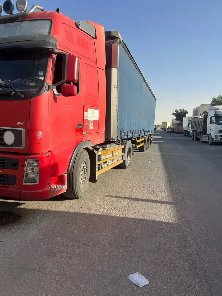 شاحنة نقل كبيرة باللون الأحمر على جانب الطريق، وتظهر عدة شاحنات أخرى تابعات لمكتب تاجير تريلات الرياض
